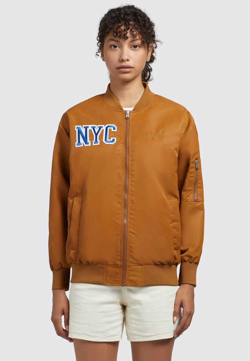 Giubbotto bomber marrone con polsini a costine, chiusura frontale con zip, ricamo NYC in blu e bianco sul petto, e tasche laterali. Tessuto di superficie morbida.