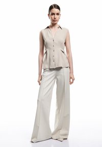 Beige ärmellose Bluse mit Knöpfen vorne und Seitenausschnitten. Kombiniert mit hoch taillierten, weit geschnittenen Hose aus passendem beigem Stoff. Glatte Textur.
