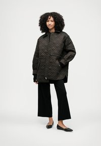 Πουπουλένιο μαύρο μπουφάν με ψηλό γιακά, μεγάλες τσέπες και πλάγιες σχισμές. Συνδυασμένο με μαύρο παντελόνι φάρδων και επίπεδες πατούσες.