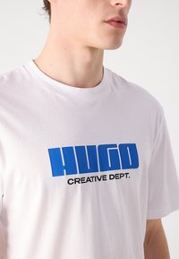 Camiseta de algodón blanca con un logo azul de "HUGO" y el texto "CREATIVE DEPT." en negro. Cuello redondo y mangas cortas, textura suave.