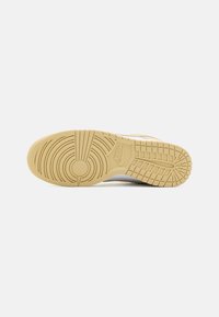 Semelle d'une basket Nike beige montrant des motifs de bande de roulement circulaires et en grille avec le logo Nike au centre sur un fond blanc uni.