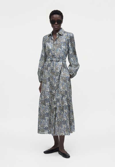 Γυναίκα με σκούρα γυαλιά ηλίου, μακρύ φλοράλ φόρεμα-πουκάμισο με φουσκωτά μανίκια και μαύρα επίπεδα παπούτσια, στέκεται μπροστά σε αδιάφορο φόντο.