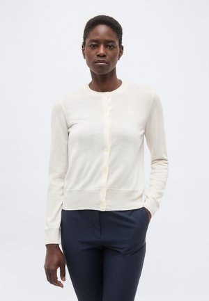 Frau trägt einen weißen, geknöpften Cardigan und dunkelblaue Hose, steht mit einer Hand in der Tasche vor einem schlichten hellen Hintergrund.