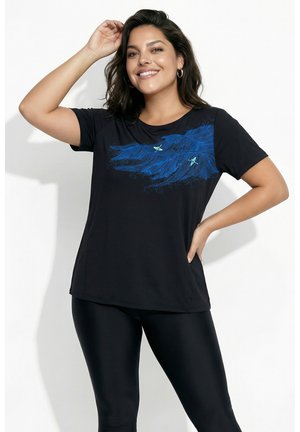 Kvinde smilende med hånd hævet, iført sort t-shirt med blåt bølge- og fugledesign og sorte leggings mod hvid baggrund.
