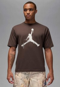 Camiseta de algodón marrón con un gran estampado blanco del logo de Jordan, mangas cortas y diseño de cuello redondo. Textura suave y ajuste relajado.