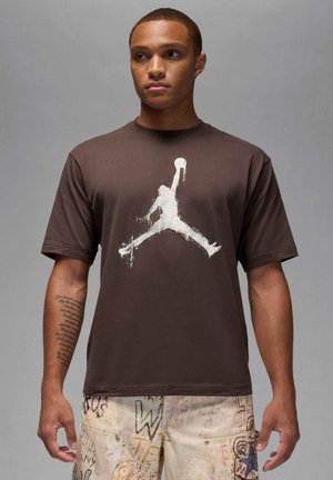 Camiseta de algodón marrón con un gran estampado blanco del logo de Jordan, mangas cortas y diseño de cuello redondo. Textura suave y ajuste relajado.