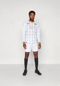 Fehér, hosszú ujjú focimez fekete geometriai mintával és arany díszítéssel, fehér nadrággal és fekete zoknival, sportos ruházat bemutatása.