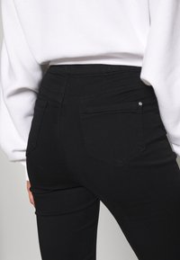 Mustat korkeavyötäröiset farkut, joissa on sileä pinta, takataskut ja pieni koristeellinen metalliyksityiskohta ylätaskun lähellä.