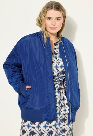 Giacca bomber blu con finitura lucida, polsini e orlo a costine, tasche laterali e chiusura con zip, abbinata a un vestito con motivo sottostante.
