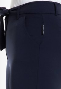 Donkerblauwe broek met een gladde textuur, voorzien van een gestrikt riem, een voorkantzak en een zichtbaar merklabel aan de zijkant.