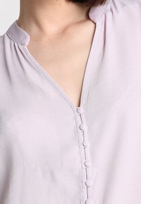Blusa lilla chiaro con scollatura a V, colletto rotondo piccolo e una patta frontale con bottoni decorativi. Tessuto liscio e strutturato.