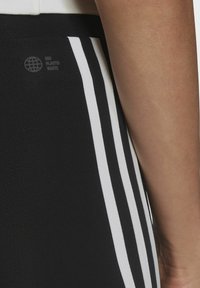 Černé sportovní kalhoty s bílými bočními pruhy a jemným logem. Materiál je hladký a těsný, navržený pro pohodlí a pohyblivost.