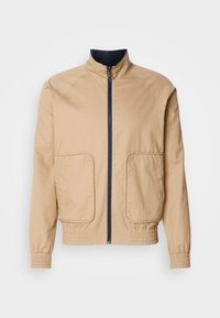 Μπεζ ελαφρύ μπουφάν με ψηλό γιακά, εμπρόσθιο φερμουάρ και δύο μεγάλες πλευρικές τσέπες. Διαθέτει ελαστικές μανσέτες και κόψιμο στο κάτω μέρος με ναυτικές λεπτομέρειες.