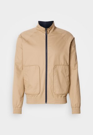 Bež lagana jakna s visokim ovratnikom, prednjim zatvaračem i dva velika bočna džepa. Ima elastične manžete i donji rub s mornarsko plavim detaljima.