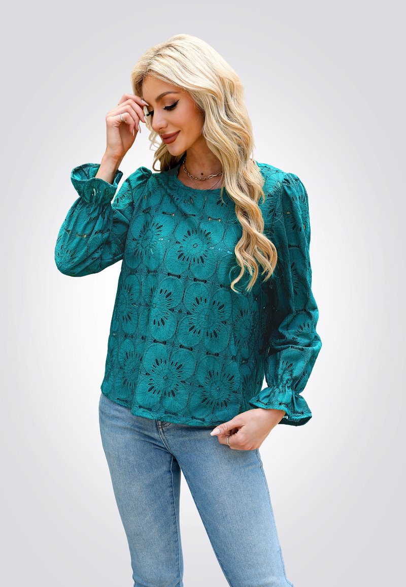 Blusa in pizzo teal con motivo floreale, scollatura rotonda e maniche a sbuffo. Abbinata a jeans azzurri chiari, dal taglio comodo.