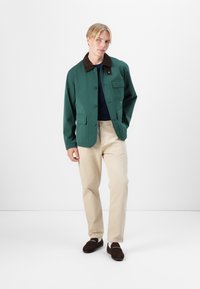 Grøn jakke med brun krave, flere frontlommer, knaplukning, båret over en marineblå top, kombineret med beige bukser og mørke loafers.