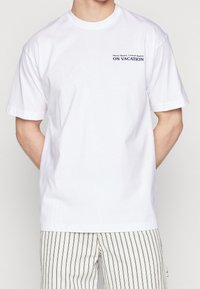 Biała koszulka z krótkim rękawem z małym napisem "Słoneczna kąpiel, koktajle na wakacjach" na górnym lewym klatce piersiowej, zestawiona z paskowanymi szortami.