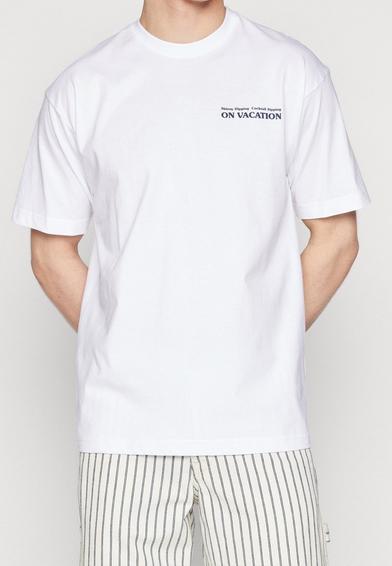 Biała koszulka z krótkim rękawem z małym napisem "Słoneczna kąpiel, koktajle na wakacjach" na górnym lewym klatce piersiowej, zestawiona z paskowanymi szortami.