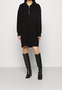 Musta pitkähihainen vetoketjupaita, jossa on rento malli, valmistettu sileästä kankaasta, yhdistettynä polvenkorkuisiin mustiin nahkasaappaisiin, joissa on korkea korko.