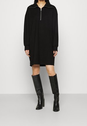 Musta pitkähihainen vetoketjupaita, jossa on rento malli, valmistettu sileästä kankaasta, yhdistettynä polvenkorkuisiin mustiin nahkasaappaisiin, joissa on korkea korko.