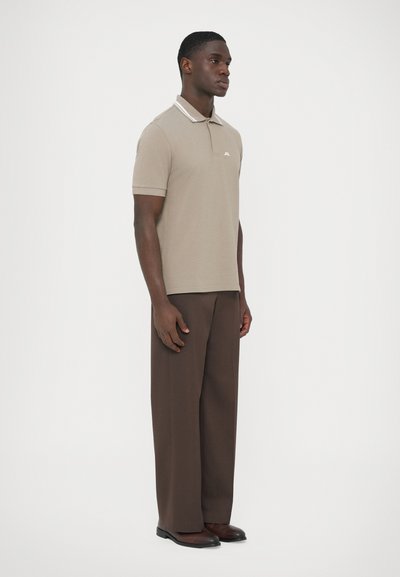 Μπεζ πόλο μπλούζα με αντιθετικό ραβδωτό γιακά, σετ με καφέ παντελόνι φαρδιάς γραμμής και σκοτεινά παπούτσια· απλό σχέδιο, κοντά μανίκια και διακριτικό λογότυπο.