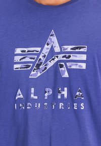 Sinine puuvillane T-särk, millel on valge ja lilla kamuflaažimustriga logo, all on rasvastes, kindlates valgetes tähtedes kirjas "ALPHA INDUSTRIES".