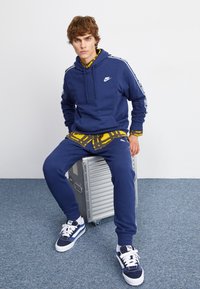 Jūras spēku Nike kapuce ar priekšējo kabatu un baltu logo, apvienota ar rakstainu dzeltenu kreklu, jūras spēku sporta biksēm un jūras spēku sporta apaviem ar baltām akcentiem.