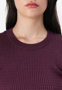 Mauve, bordás kötött felső kerek nyakkal, textúrált mintával. Az anyag puhanak és nyújthatónak tűnik, laza szabással.