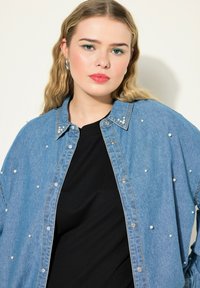 Camicia in denim con accenti di perle, colore azzurro chiaro, design con bottoni, texture morbida, indossata sopra un top nero, con colletto classico.