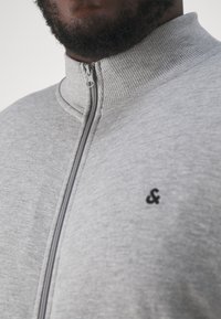 Chaqueta gris con cremallera y cuello alto, textura acanalada y logo negro de "&". Cuenta con una cremallera frontal y una tela suave y lisa.