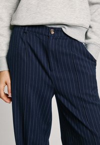 Pantalones a rayas azul marino hechos de un tejido texturizado, con un cierre de un solo botón y pliegues, combinados con una sudadera gris.