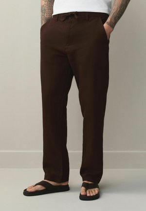 Mann trägt dunkelbraune Kordelzug-Hosen und schwarze Flip-Flops, steht mit Händen in den Taschen vor einem einfarbigen Hintergrund.