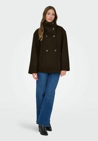 Abrigo de lana marrón oscuro, de doble botonadura, con un cuello alto y dos bolsillos laterales, combinado con jeans acampanados azules y botines negros.