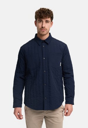 Mand iført en knappet, mørkeblå quiltet langærmet skjorte med to brystlommer, kombineret med beige bukser, stående mod en ensfarvet baggrund.