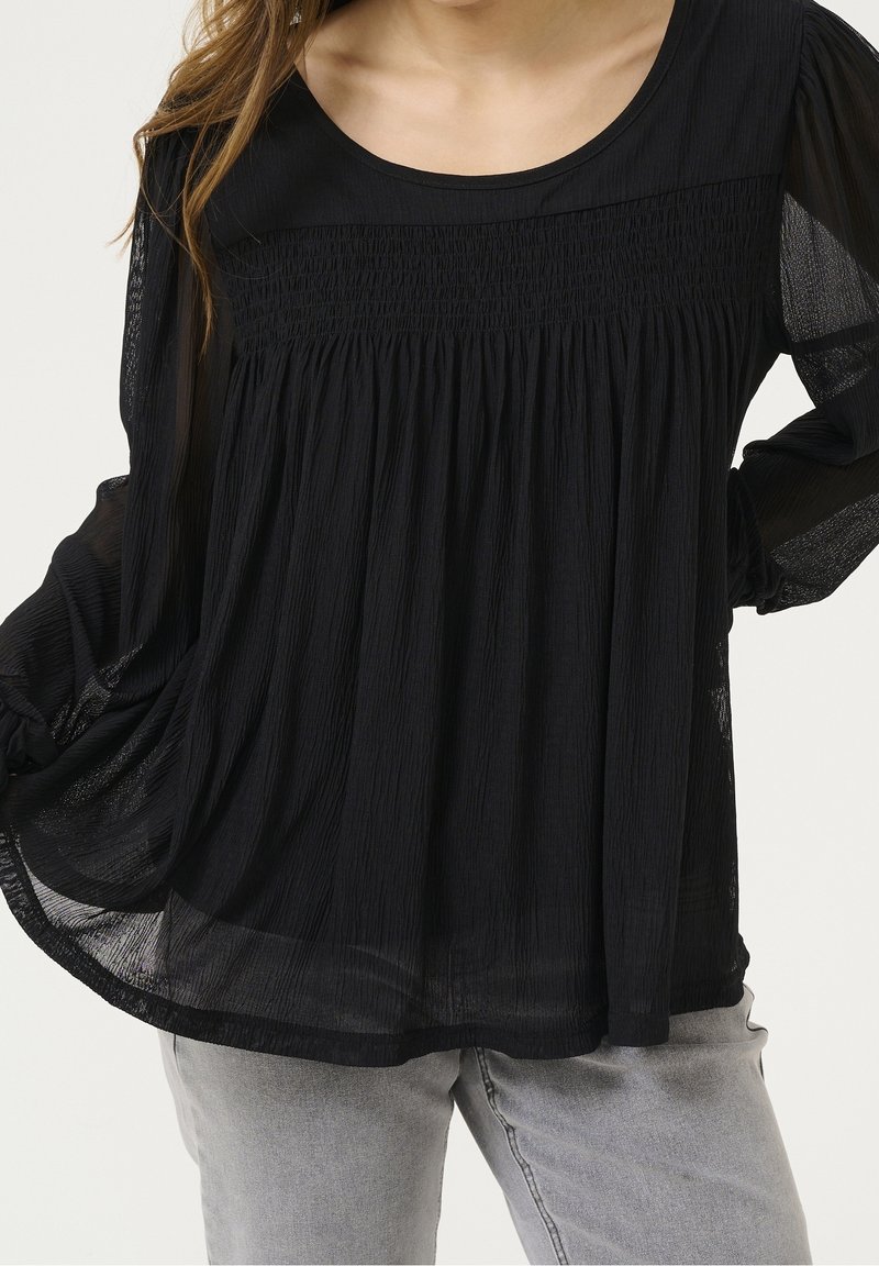 Blouse noire légère avec encolure ronde et design plissé. Comprend des manches longues semi-transparentes et une coupe décontractée, associée à un jean gris.