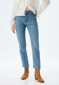 Jeans de mezclilla azul claro con corte recto, cintura alta, diseño de cinco bolsillos y detalles sutiles de decoloración, combinados con botas de tobillo color tostado.