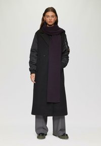 Manteau noir long avec des manches en tissu noir contrastant, associé à une écharpe violette foncée, un pantalon large gris et des chaussures noires chunky.
