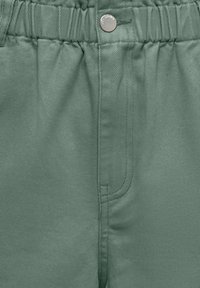Gros plan d’un pantalon vert avec ceinture élastique, bouton métal et coutures visibles sur la poche avant.