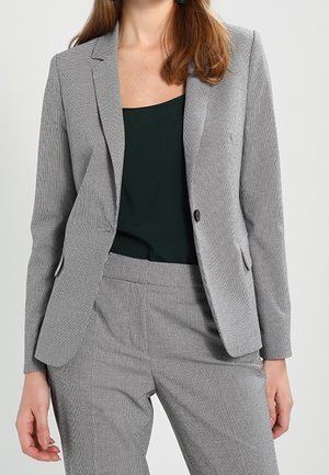 Kvinde iført gråternet skræddersyet blazer og matchende bukser over en mørkegrøn ærmeløs top.