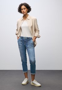 Camicia button-up beige chiaro sopra una t-shirt bianca. Jeans in denim blu con una vestibilità rilassata, orli arrotolati e leggermente logori. Sneakers bianche.