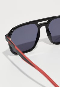 Ochelari de soare rectangulari negri cu lentile întunecate, având un design cu punte despărțită și temple cu accente roșii pentru un aspect modern.