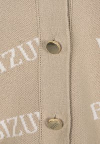 Beige gebreide cardigan met witte tekst, ronde halslijn en twee goudkleurige knopen. Zachte textuur met een casual ontwerp.