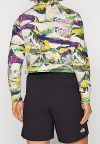 Mångfärgad fleecetröja med bergstryck, hög krage och logotyp på ryggen. Matchad med svarta shorts och synlig sidoficka.