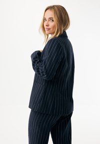 Marynarka w granatowe paski z kołnierzem, o luźnym kroju i bocznych kieszeniach. Teksturowana tkanina z subtelnymi metalicznymi nitkami biegnącymi pionowo.