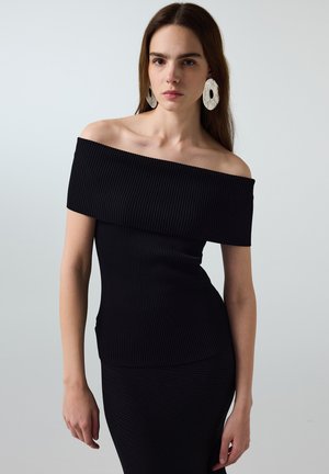 Donna che indossa un vestito nero aderente a coste, scollato sulle spalle, e grandi orecchini ovali bianchi con texture su sfondo grigio uniforme.