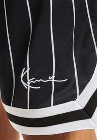 Černé sportovní kraťasy s bílými vertikálními pruhy, které mají smyčkovou strukturu a kontrastní bílý a černý pruhovaný lem. Přítomný podpisový detail.