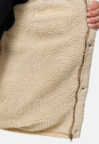 Beige szövet mellény texturált felülettel, arany cipzárral és bordázott aljjal. Tartalmaz egy kis belső zsebet a tetején.
