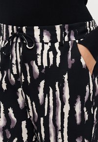 Černobílé abstraktní vzorované kalhoty s pružným pasem, nošeny s černým topem, ruka umístěna v pravé kapse.