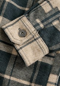 Beige und dunkelgrünes Karoshemd mit Knopfverschluss am Ärmel. Der Stoff hat eine weiche Textur und weist auffällige Karomuster sowie kontrastierende Farben auf.
