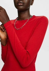 Red long-sleeve top with a scalloped neckline and fitted sleeves, paired with a silver chain necklace featuring circular links.